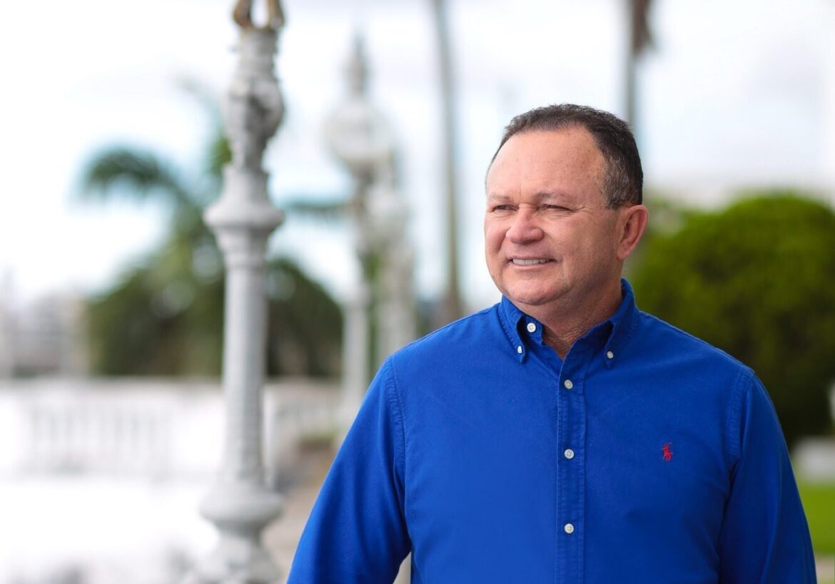 Homem de meia-idade vestindo camisa azul royal de mangas dobradas, sorrindo levemente enquanto observa o horizonte. Ao fundo, arquitetura histórica com elementos decorativos brancos e paisagem verde desfocada.