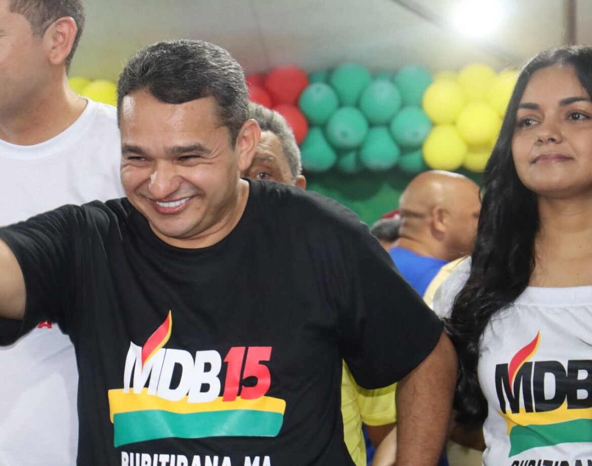 Homem de camiseta preta com logotipo do MDB15 sorri e faz sinal positivo com a mão direita em evento político decorado com balões vermelhos, verdes e amarelos, com pessoas ao redor