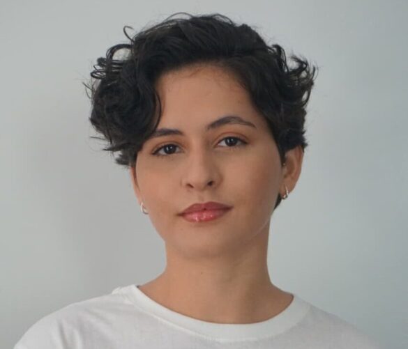 Foto de Kethlen Mata, repórter do Atual7, mulher de cabelos cacheados curtos e escuros, usando camiseta branca com estampa floral colorida, em fundo cinza claro.